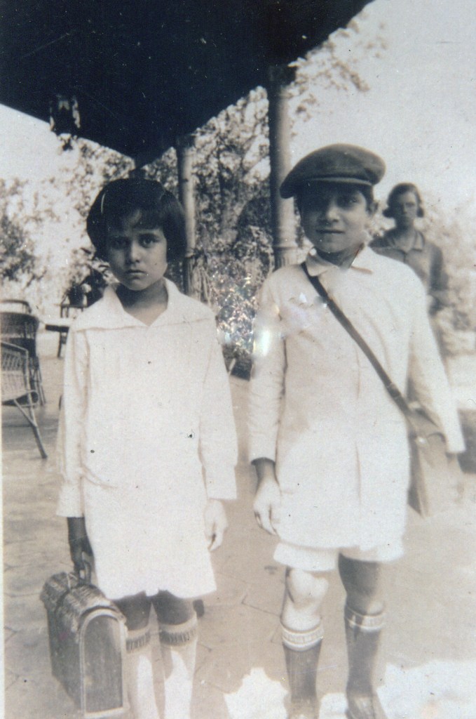 Niño Augusto Roa Bastos, con guardapolvos de la escuela, de gorra y portafolio cruzado al pecho.  Su hermana de guardapolvos y portafolio en la mano derecha; posan en la galería de la casa